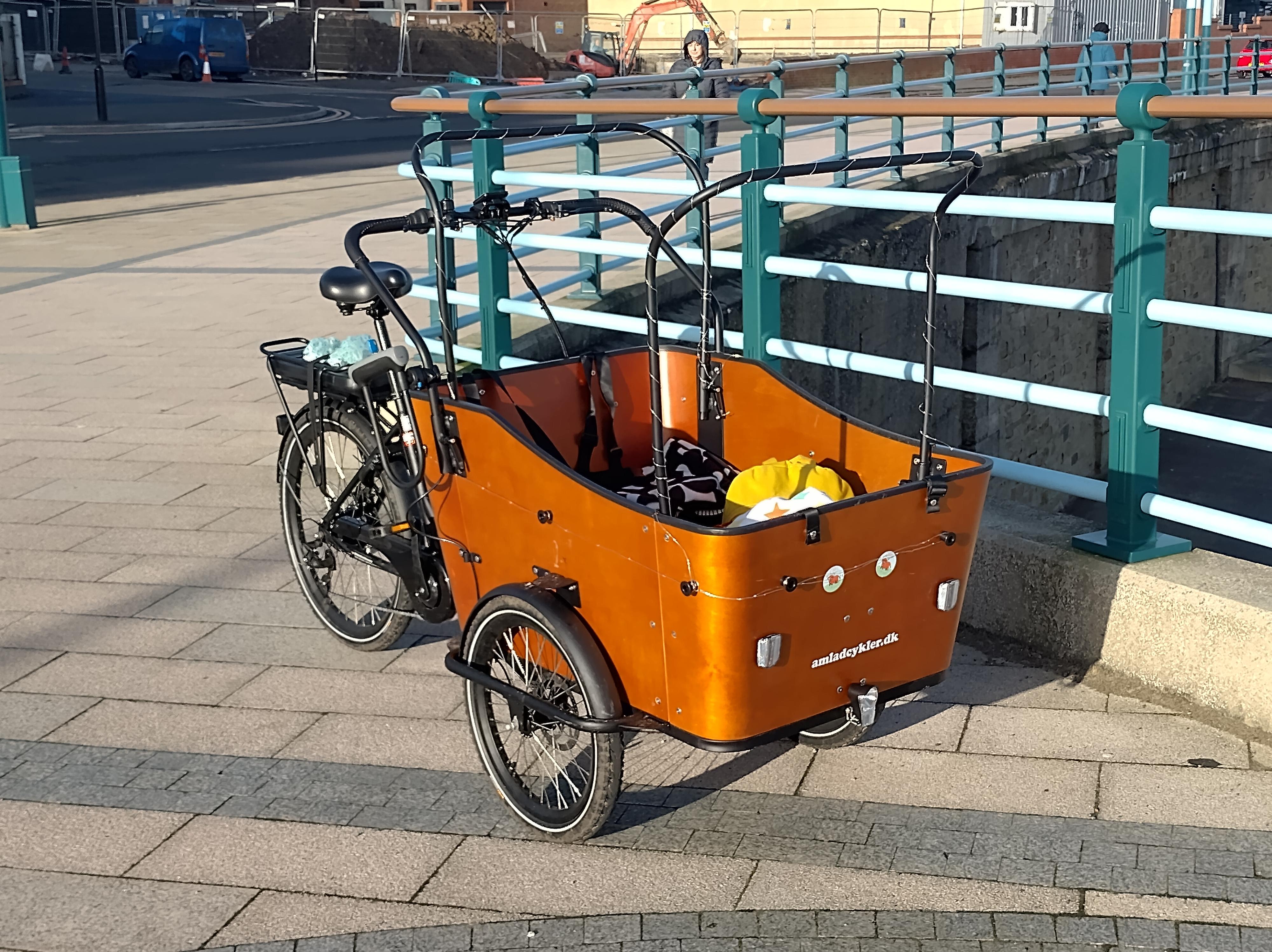 The cargo bike standing on the beach front. It has a large wooden bucket on the front, supported by two wheels. There are poles for the roof, and a handlebar like a pram handle. The back half is like a normal bike.