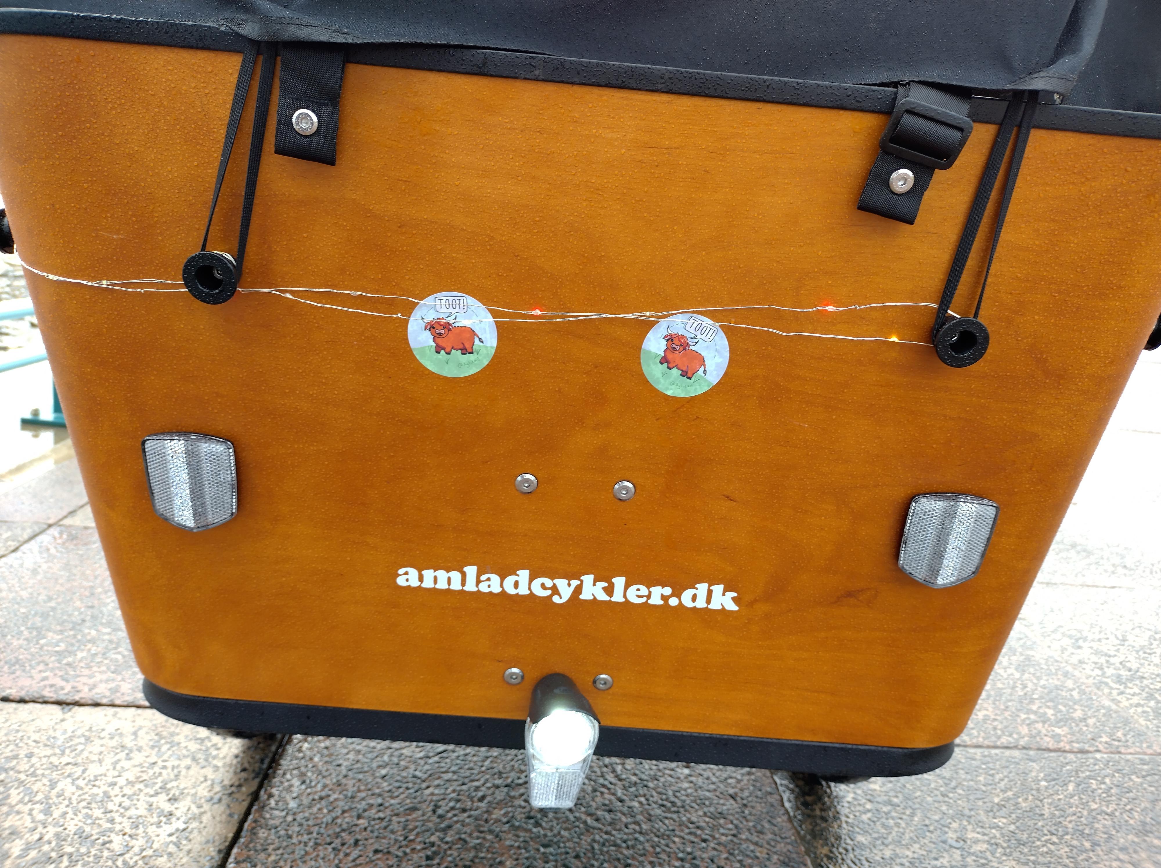 The front of the bucket. Two circular stickers above a white amladcykler.dk logo. There are two reflectors on the outer edges. The front light is under the logo.