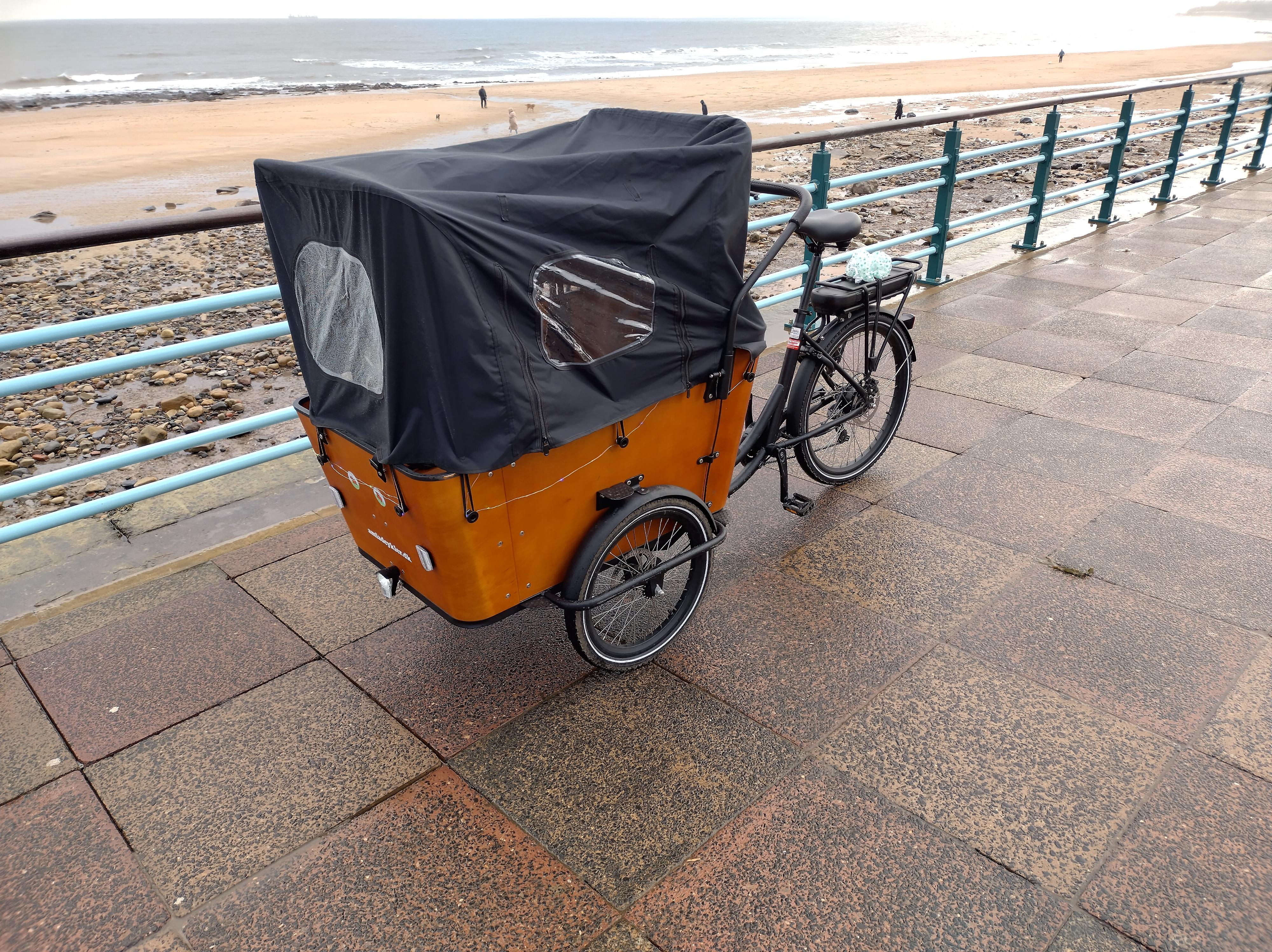 The bike viewed from the side, with the rain cover on. The cover is big and black, with transparent plastic windows on each side.